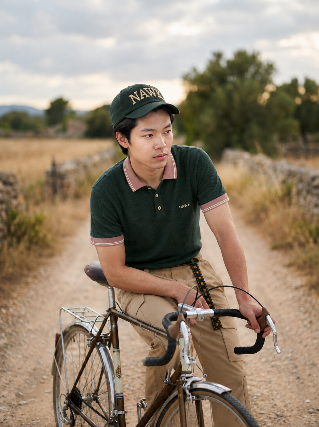 Man in a NAWK cap resting on a bicycle in the countryside